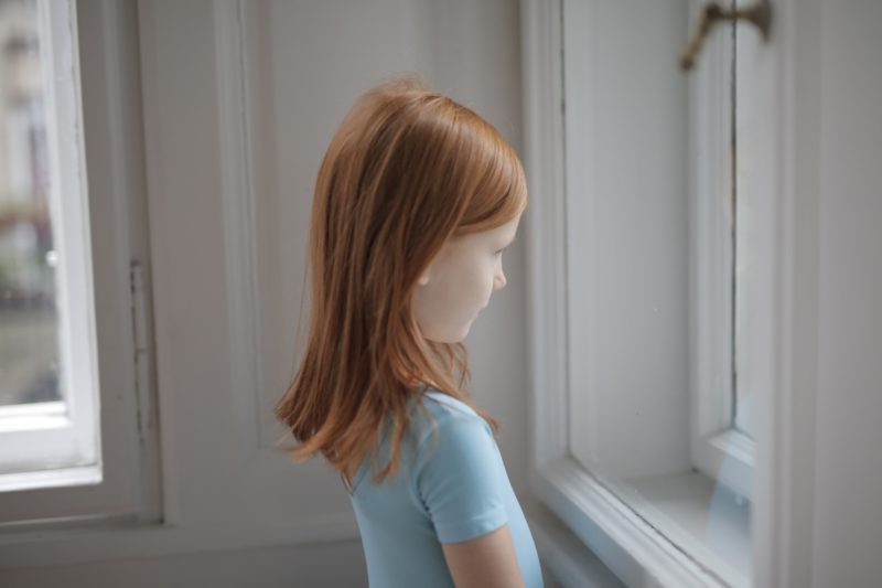 small girl with red hair and blue shirt looking out window